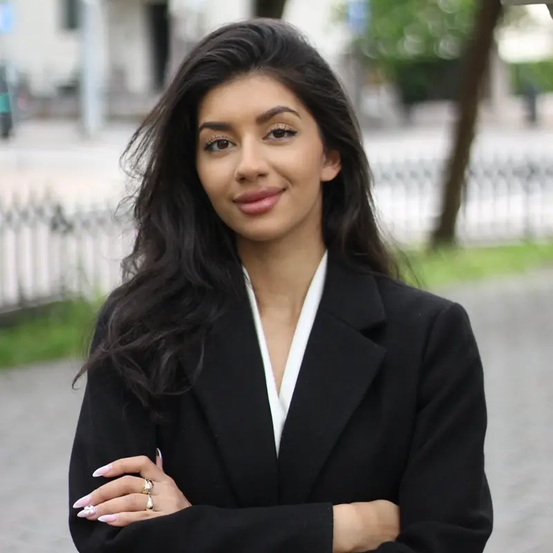 En kvinne med langt, mørkt hår, iført svart blazer og hvit topp, står utendørs med armene i kors og et lite smil mot kameraet - hun representerer den profesjonelle ånden til Om Onsite Technologies.