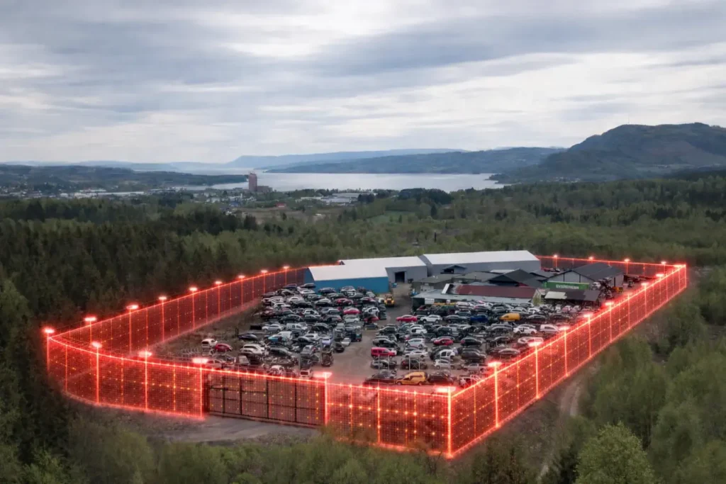 På en stor inngjerdet tomt fylt med parkerte biler, omgitt av røde lys, ligger Onsite-løsningen for bildemonteringsbransjen i et landlig område med åser og vann i bakgrunnen.